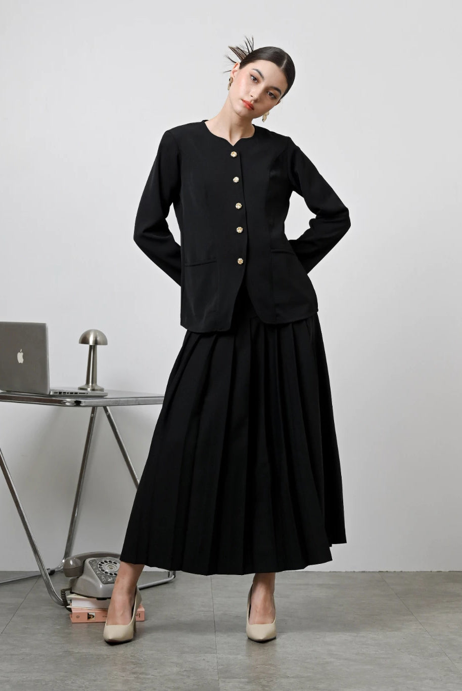 A model wearing a black Zora skirt by Kasa Heritage with pleats, paired with a matching brown top. The skirt reaches mid-calf and has a fitted waist with a zipper at the back.
