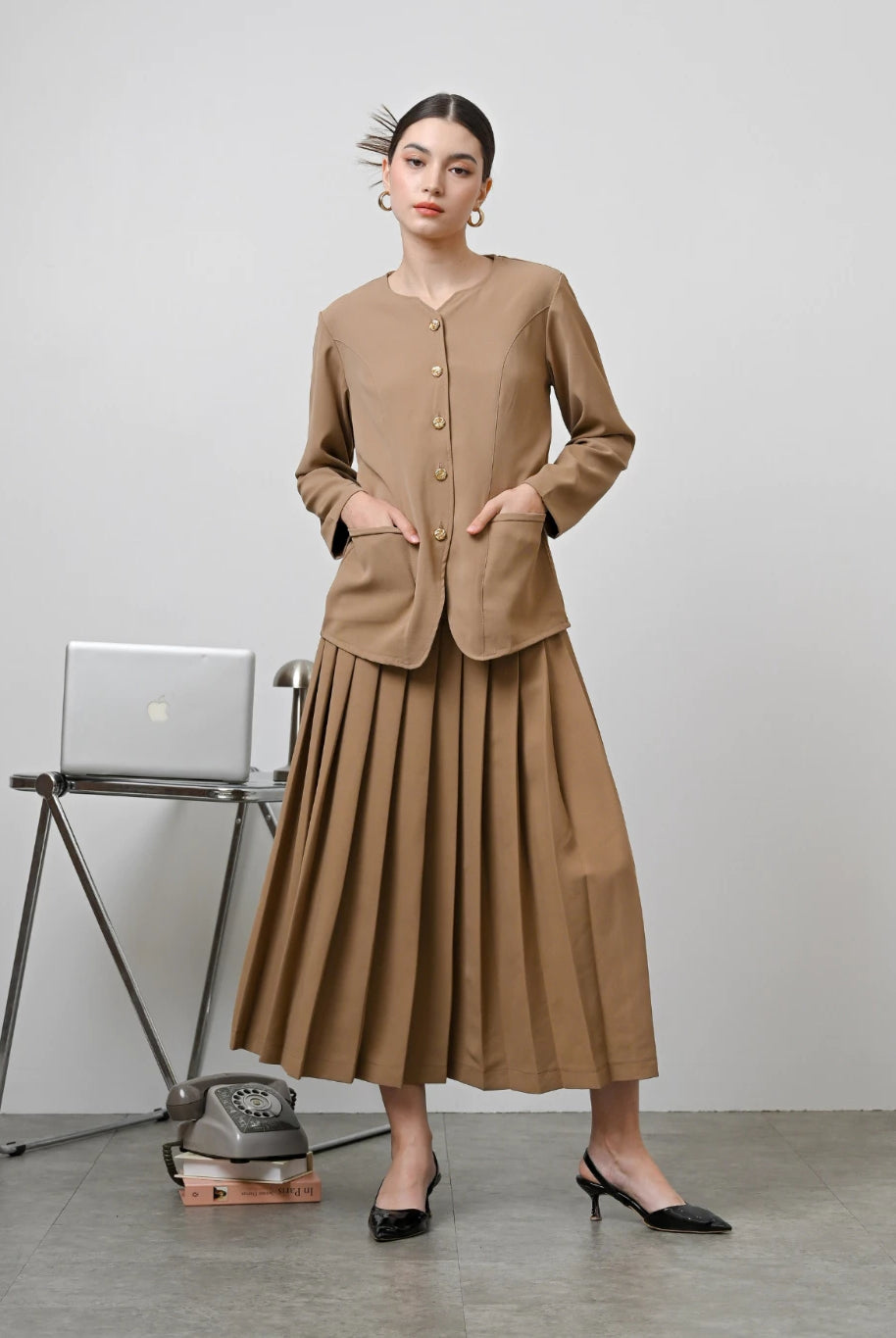 A model wearing a brown Zora skirt by Kasa Heritage with pleats, paired with a matching brown top. The skirt reaches mid-calf and has a fitted waist with a zipper at the back.