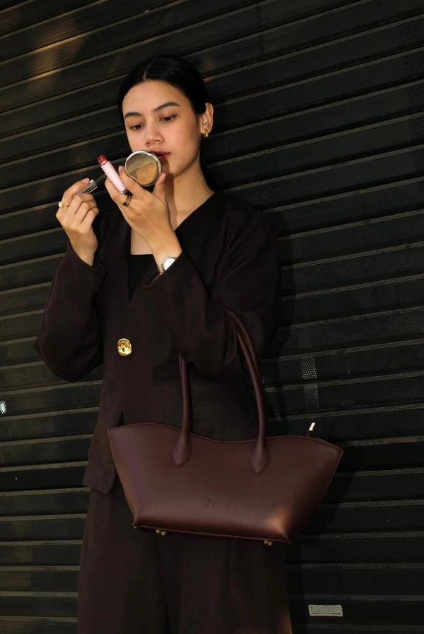 Woman in a brown outfit holding a burgundy woman's handbag by Prior against a building background.
Seorang wanita berpakaian cokelat memegang tas tangan wanita berwarna burgundy dari merek Prior di depan latar belakang bangunan.