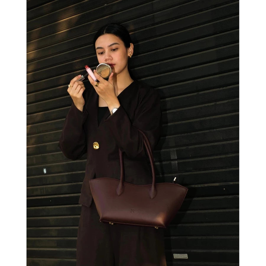 Woman in a brown outfit holding a burgundy woman's handbag by Prior against a building background.
Seorang wanita berpakaian cokelat memegang tas tangan wanita berwarna burgundy dari merek Prior di depan latar belakang bangunan.