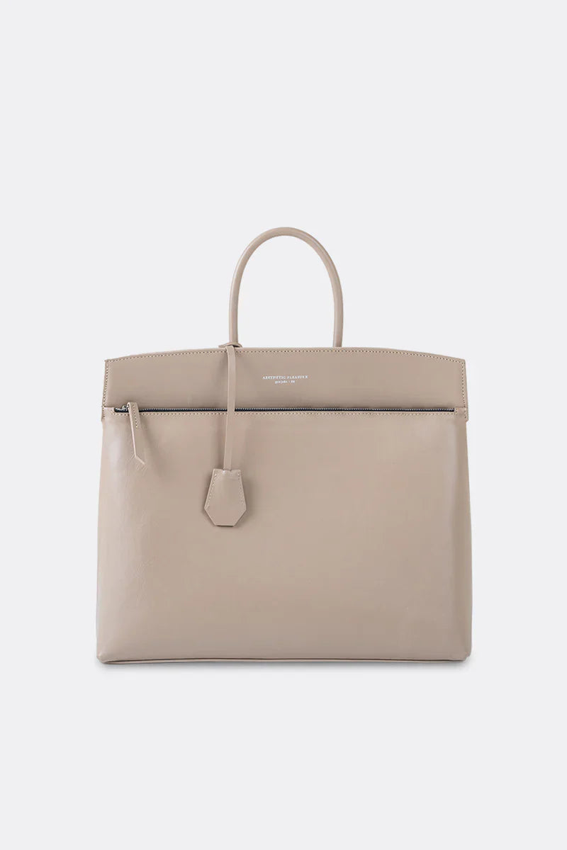 A beige nappa leather handbag with a structured design, featuring a tubular handle, a logo stamp in front, and a detachable hanging leather key fob by Aesthetic Pleasure.