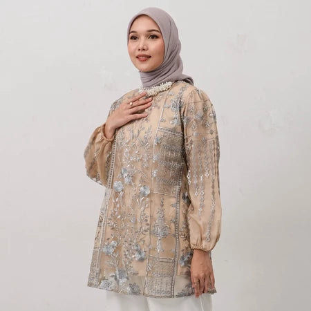A woman wearing a beige tunic with floral embroidery and long sleeves, paired with white pants and a grey headscarf.