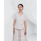 A woman wearing a beige, asymmetrical button-down blouse with short sleeves and a round neckline by Kasyalia.