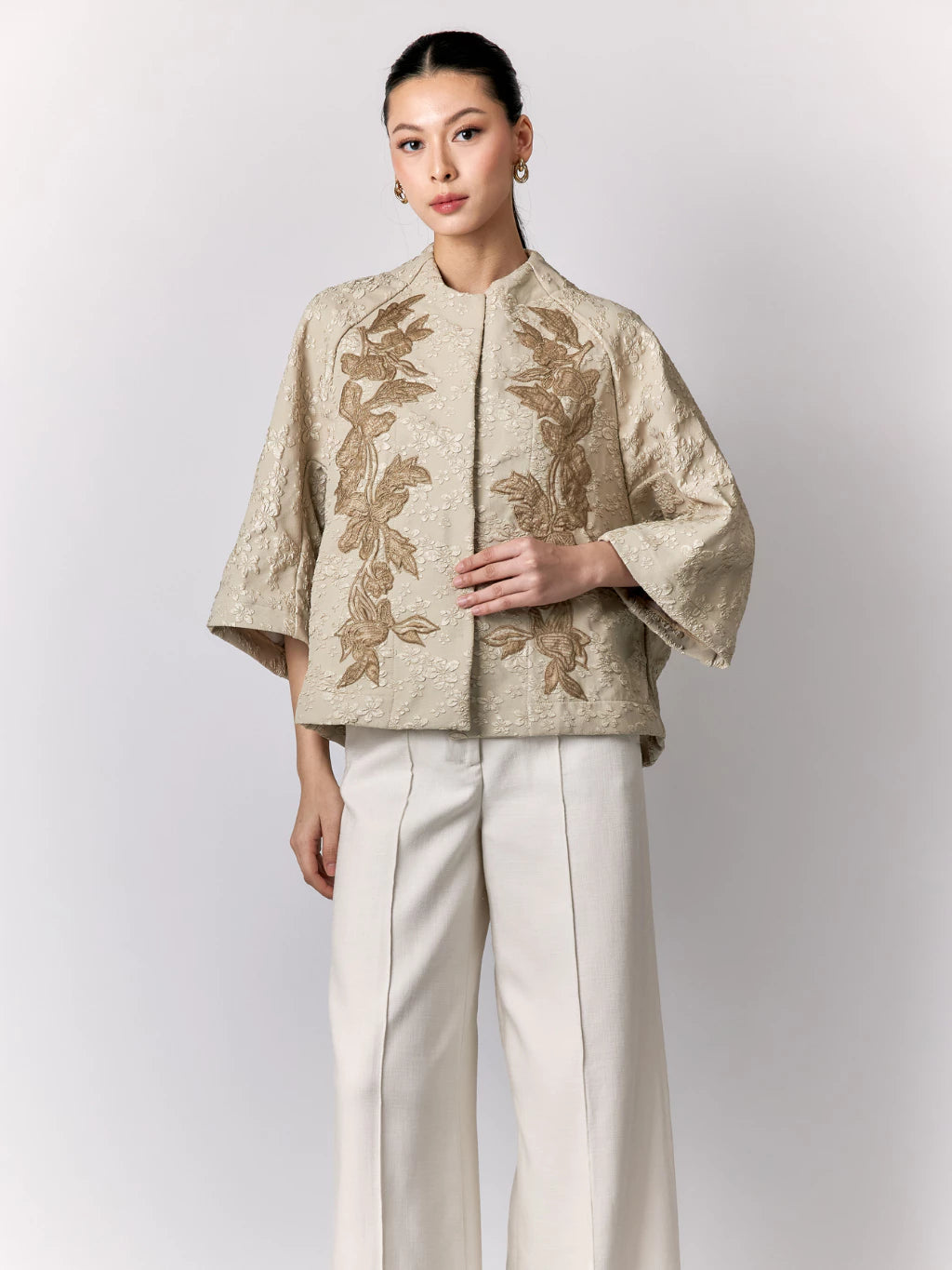 Woman wearing a beige embroidered jacket by Studio Tui and white pants on a light gray background