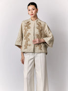 Woman wearing a beige embroidered jacket by Studio Tui and white pants on a light gray background