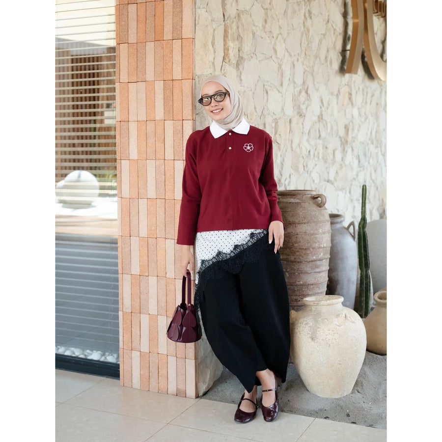 A woman wearing a maroon long-sleeve polo shirt by Haidee Orlin with a white collar, black wide-leg pants, and a beige hijab stands in an outdoor patio setting next to a tiled column.