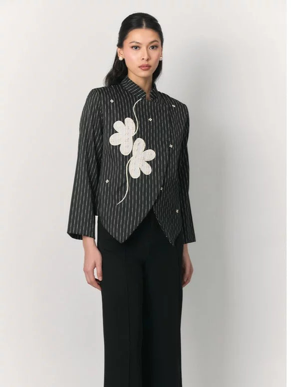 A model wearing a black and white vertically striped top by Studio Tui with a large white embroidered flower on the chest, paired with black trousers, against a plain light gray background.
