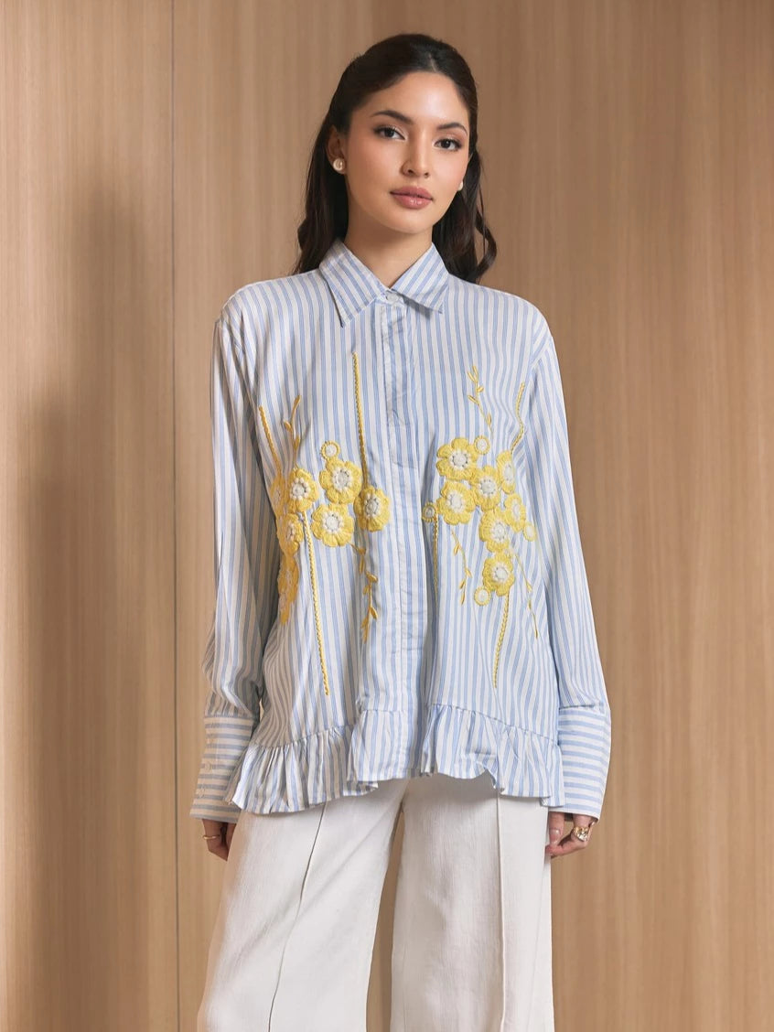 A woman models a light blue and white vertically striped long-sleeve shirt by Studio Tui with yellow floral embroidery on the front, paired with white trousers, set against a warm wood-paneled background.