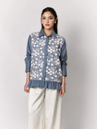 A woman models a slate blue button-up shirt by Studio Tui with a white floral embroidered pattern over the front panel, paired with cream wide-leg trousers, against a light gray studio background.
