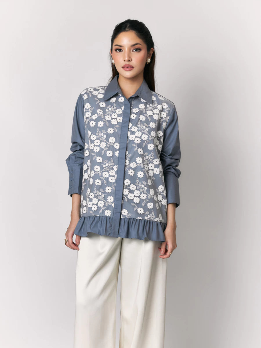 A woman models a slate blue button-up shirt by Studio Tui with a white floral embroidered pattern over the front panel, paired with cream wide-leg trousers, against a light gray studio background.