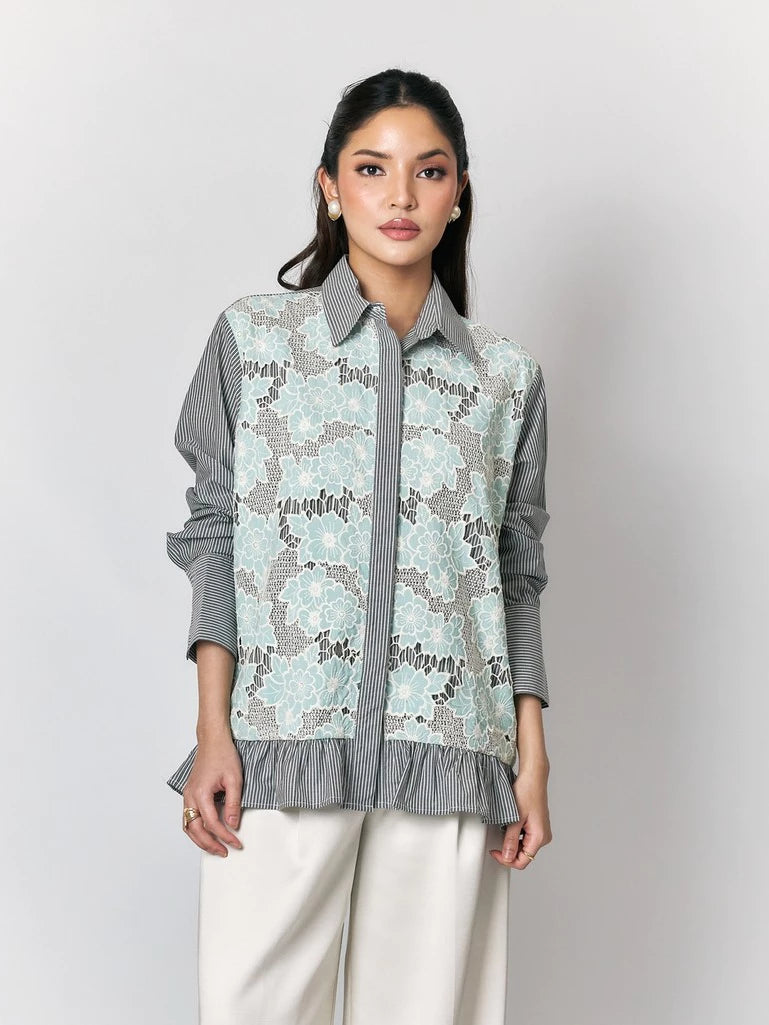 A woman models a long-sleeve button-up shirt by Studio Tui with a light blue and gray floral lace overlay on a gray striped base, paired with white trousers, against a neutral gray background.