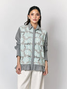 A woman models a long-sleeve button-up shirt by Studio Tui with a light blue and gray floral lace overlay on a gray striped base, paired with white trousers, against a neutral gray background.