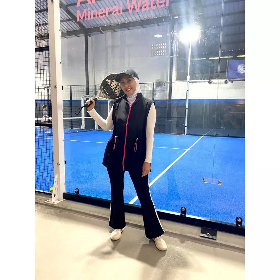 Woman wearing a modest sporty outfit featuring a black sleeveless vest by Abame with zipper details, a light-colored long-sleeve inner top, black flare pants, and white sneakers at an indoor padel court.