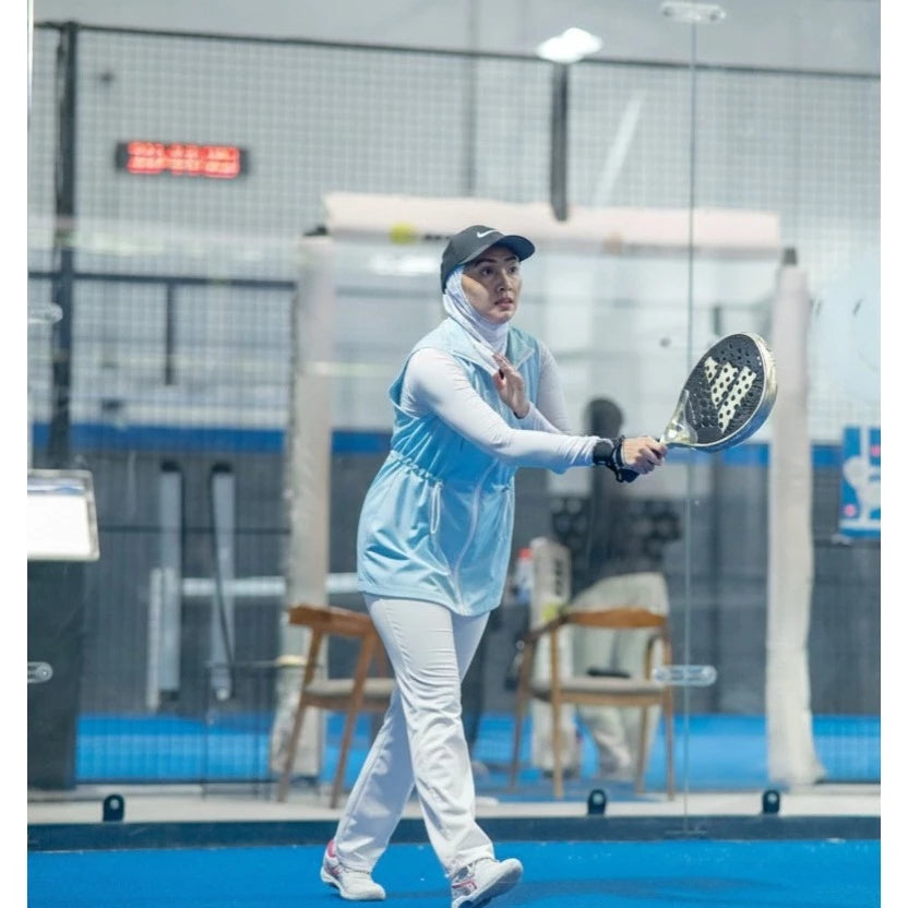 Woman wearing a modest sporty outfit featuring a light blue sleeveless vest by Abame with zipper details, a light-colored long-sleeve inner top, white flare pants, and white sneakers at an indoor padel court.