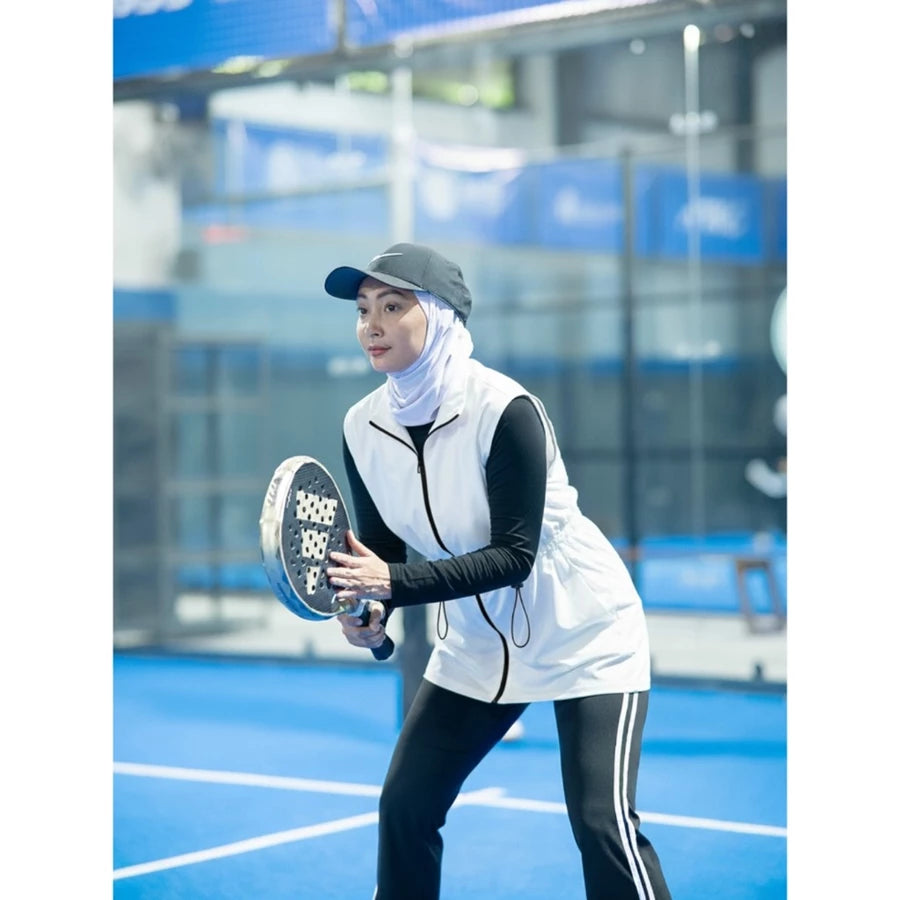Woman wearing a modest sporty outfit featuring a broken white sleeveless vest by Abame with zipper details, a light-colored long-sleeve inner top, and black flare pants at an indoor padel court.