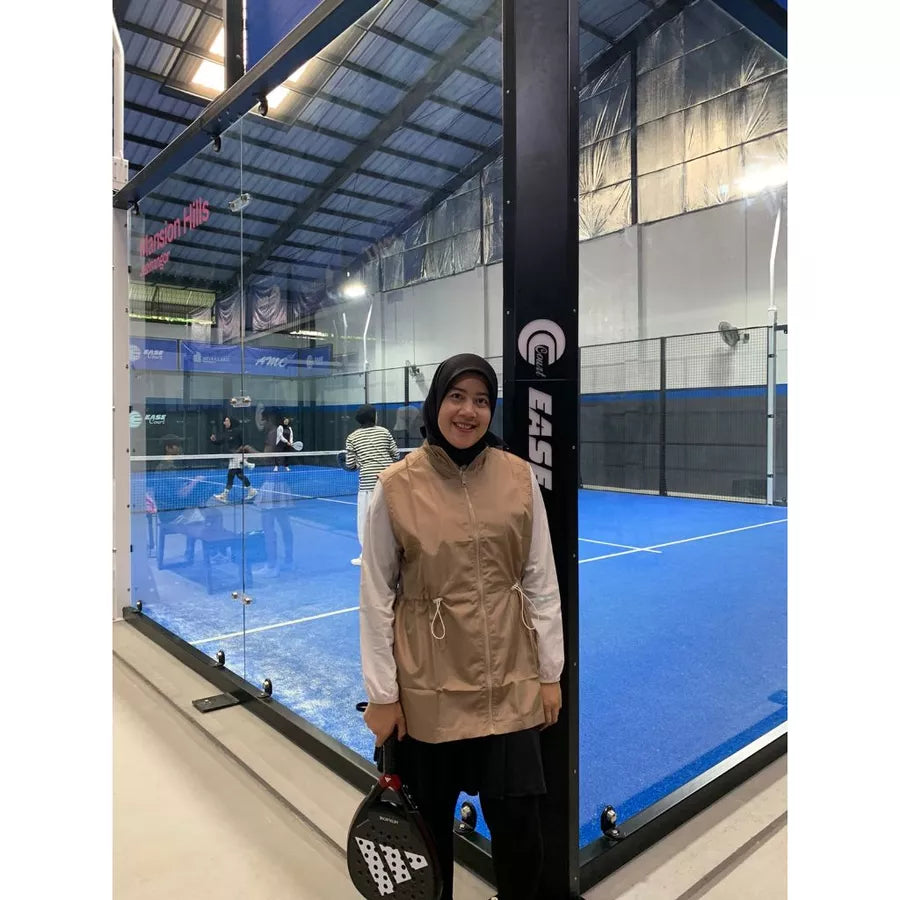 Woman wearing a modest sporty outfit featuring a light brown sleeveless vest by Abame with zipper details, a light-colored long-sleeve inner top, and black flare pants at an indoor padel court.