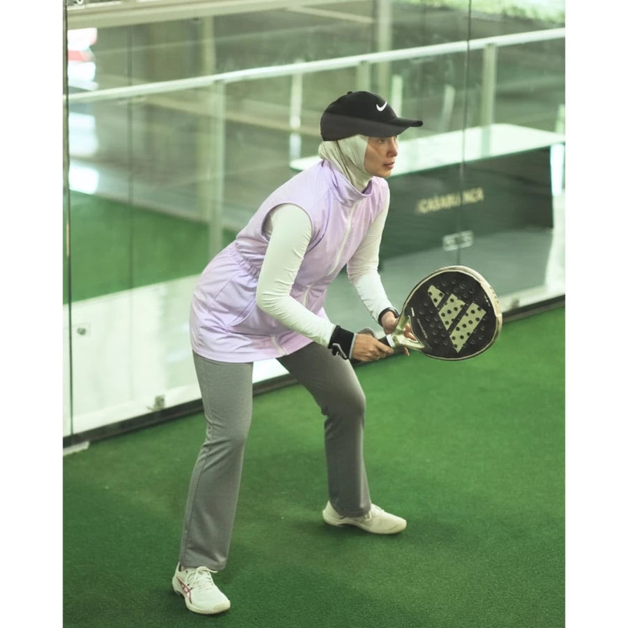 Woman wearing a modest sporty outfit featuring a lilac-colored sleeveless vest by Abame with zipper details, a light-colored long-sleeve inner top, light grey flare pants, and white sneakers at an indoor padel court.