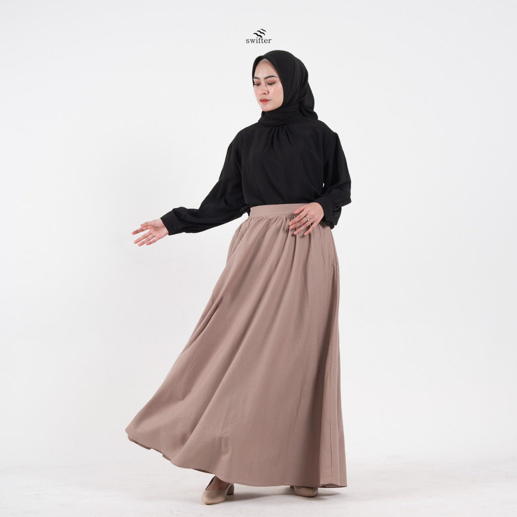 A woman wearing an ash brown linen skirt by Swifter with a wide hemline, flowing fabric, and a lining. She has a zipper at the left side and a pocket at the right side on the skirt.