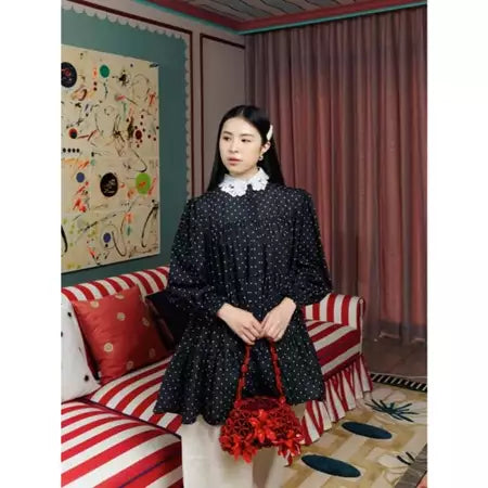 Woman in a black polka dot blouse by Haidee Orlin holding a red bag in a room with colorful walls and a striped couch.