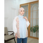 Woman wearing a light blue shirt by Abame with a floral details in a room with a wooden door and chair.