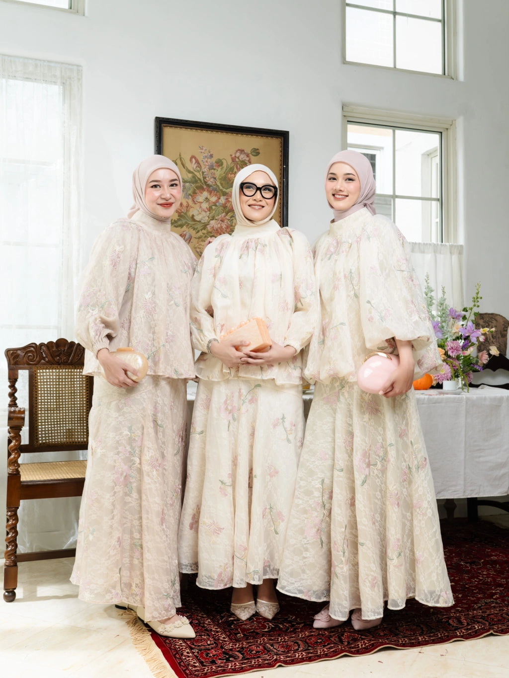 Three women in matching white outfits by Haidee Orling standing in a room with large windows and a table with flowers.