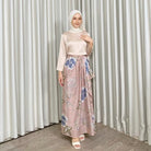 Woman wearing a floral skirt by Kartinis Label and beige outfit standing against a plain curtain background