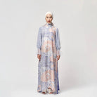 A woman standing in a studio setting, wearing a long, floral and geometric patterned dress by Kartinis Label with a denim-like texture.