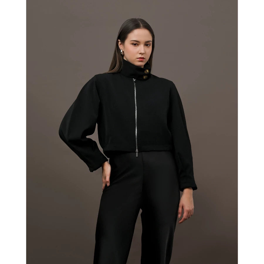 A model wearing a black cropped jacket  by Kasa Heritage with a high mock neck, silver front zipper, and decorative gold buttons near the collar, paired with black trousers, set against a solid dark brown studio background.