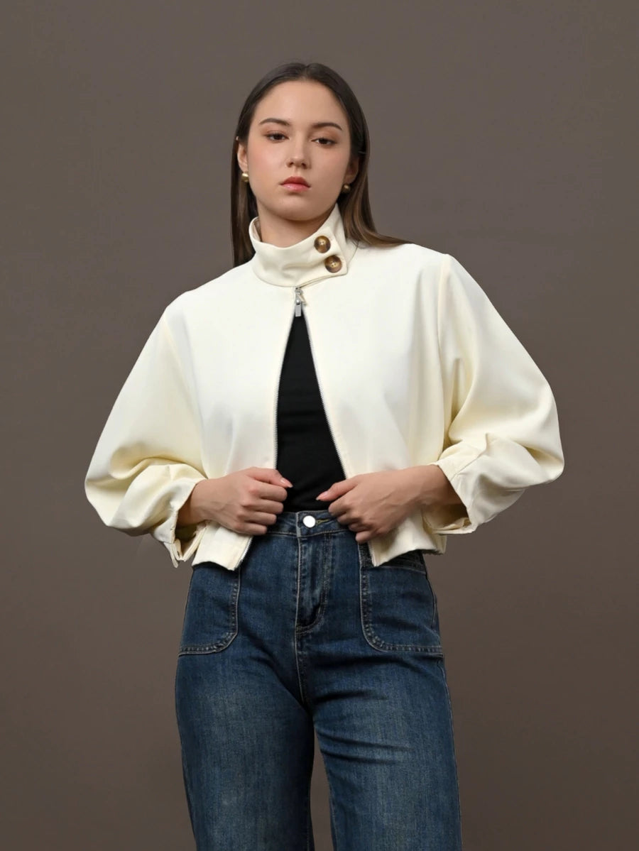 A woman modeling a cream-colored cropped jacket by Kasa Heritage with a high mock neck and gold-tone buttons, worn over a black top and blue jeans, against a solid dark gray studio background.
