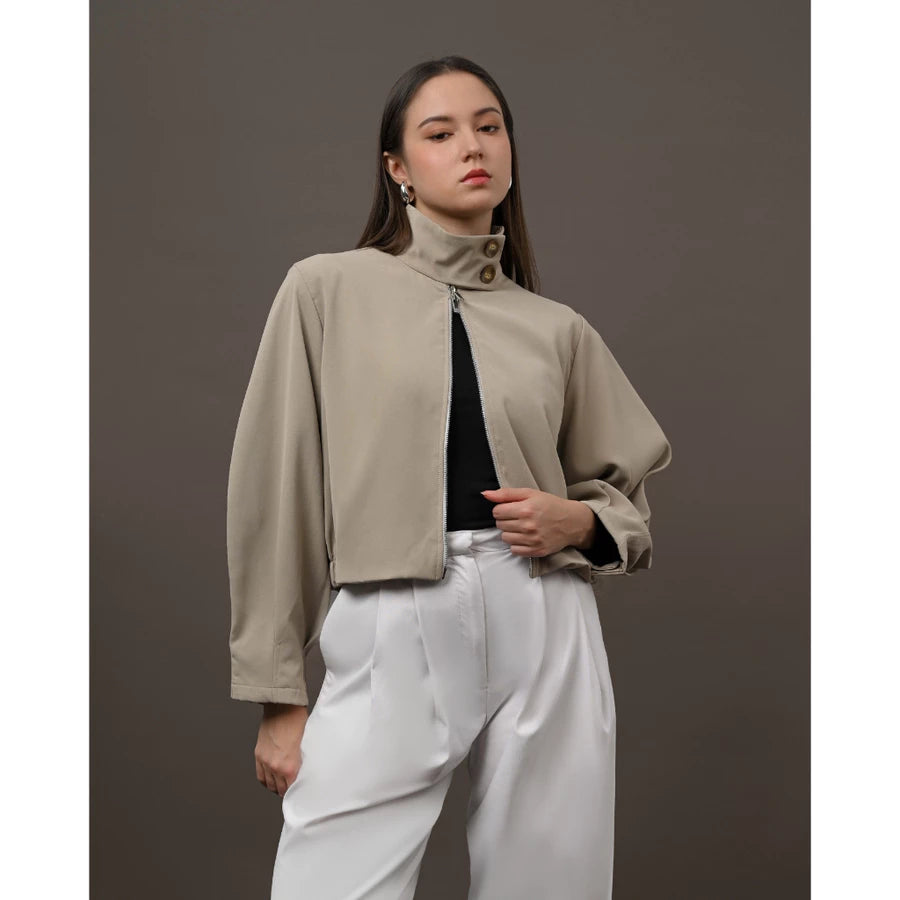 A woman modeling a cropped, high-neck beige jacket by Kasa Heritage with a front zipper, worn over a black top and white trousers, set against a solid dark gray studio background.