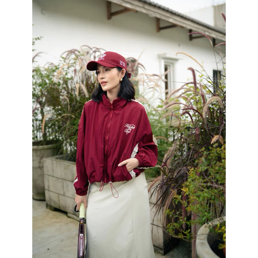 Person wearing a maroon windbreaker jacket by Haidee Orlin and cap holding a tennis racket in an outdoor setting
