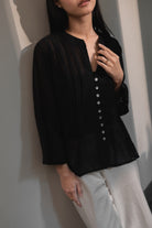 Person wearing a black button-up blouse by Kasyalia with white pants against a neutral background