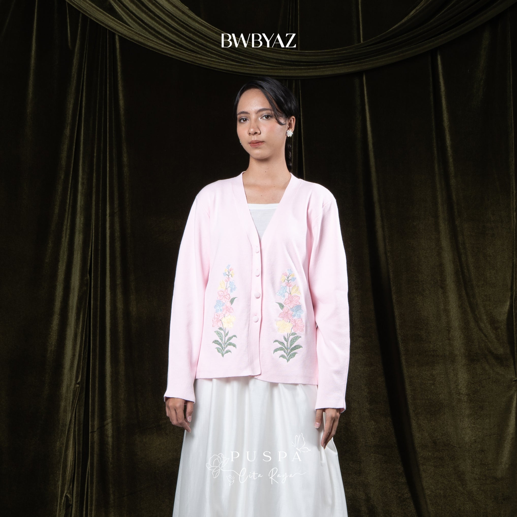 Woman wearing a pink embroidered cardigan over a white outfit against a dark curtain background with 'BWBYAZ' branding.
