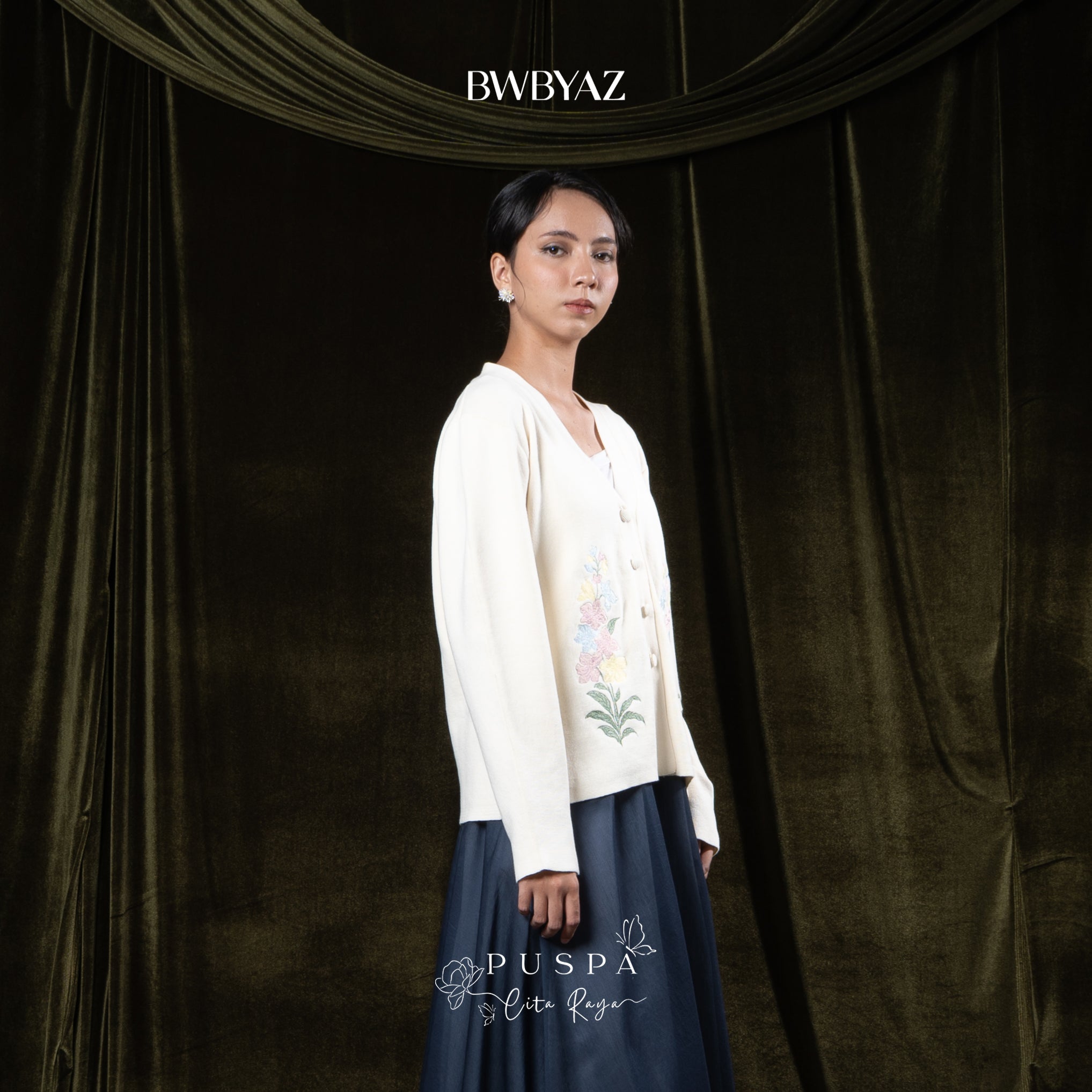 Woman wearing a cream-colored embroidered cardigan over a white outfit against a dark curtain background with 'BWBYAZ' branding.