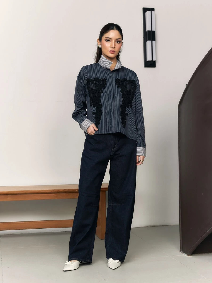 Woman wearing a navy shirt with black floral embroidery by Studio Tui and dark pants in a minimal setting.