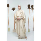 A person wearing a beige maxi dress by Glashka with long sleeves standing against a white background with decorative trees in the background.