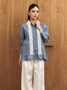 A woman models a dusty blue long-sleeve shirt by Studio Tui with white embroidered vertical panels down the front, paired with cream wide-leg trousers, standing against a wooden wall.