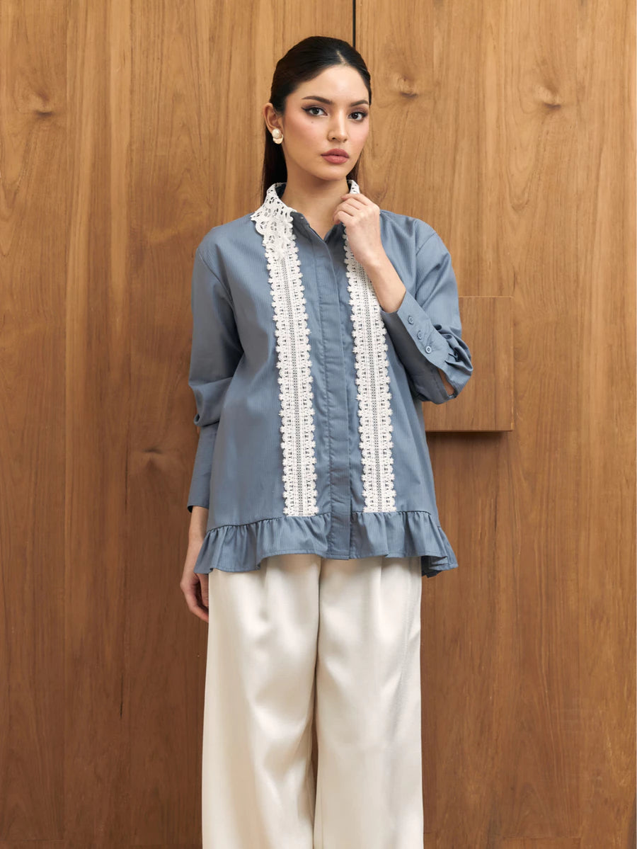 A woman models a dusty blue long-sleeve shirt by Studio Tui with white embroidered vertical panels down the front, paired with cream wide-leg trousers, standing against a wooden wall.
