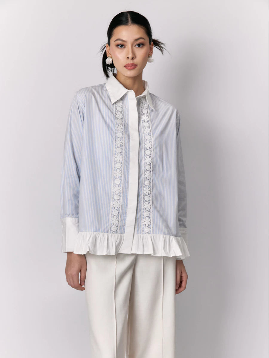 Woman wearing a light blue striped shirt by Studio Tui with white lace details and white pants on a plain background