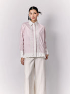 Woman wearing a light pink shirt by Studio Tui with lace details and white pants on a plain background