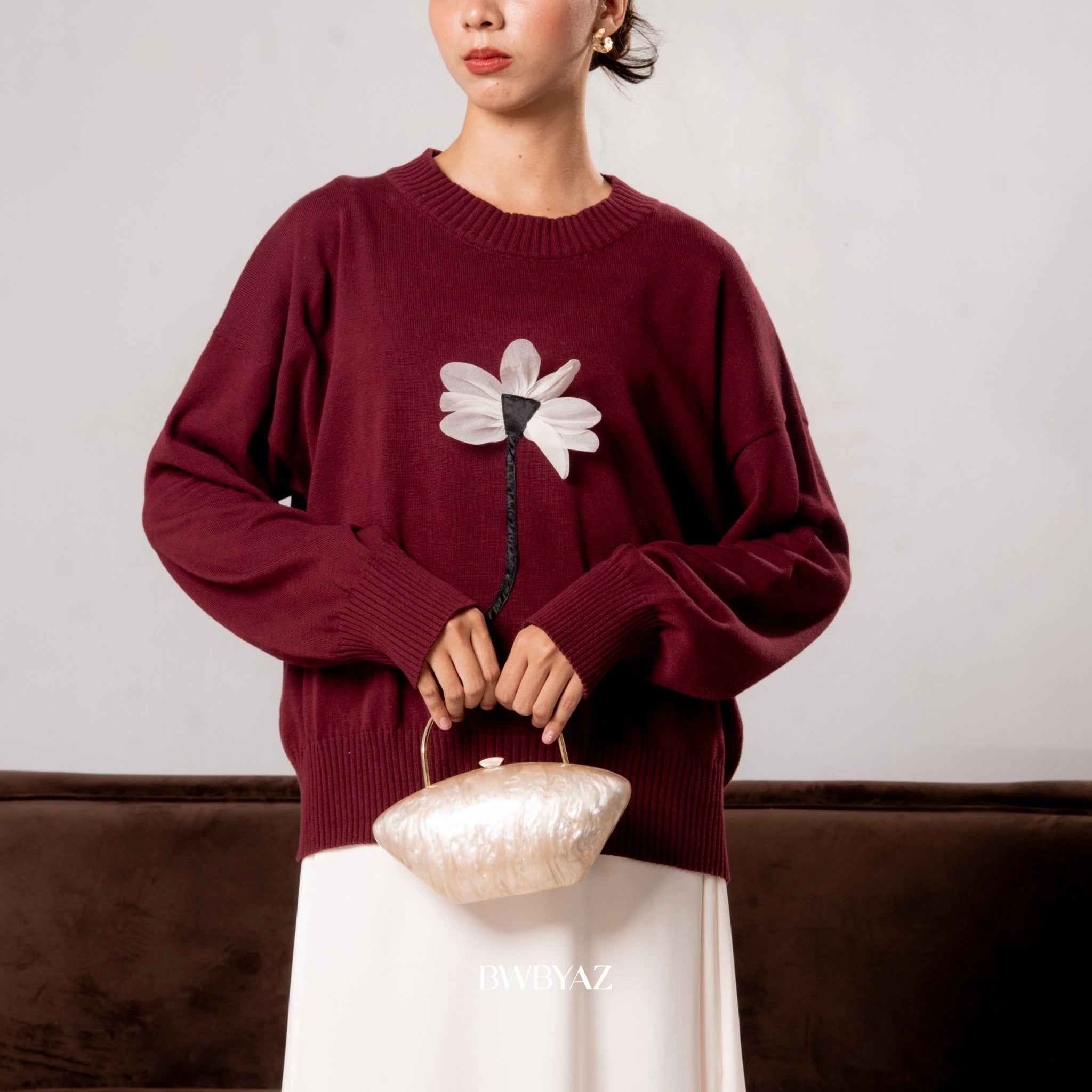 Person wearing a maroon sweater with a floral design from BWBYAZ, holding a beige handbag against a neutral background.
