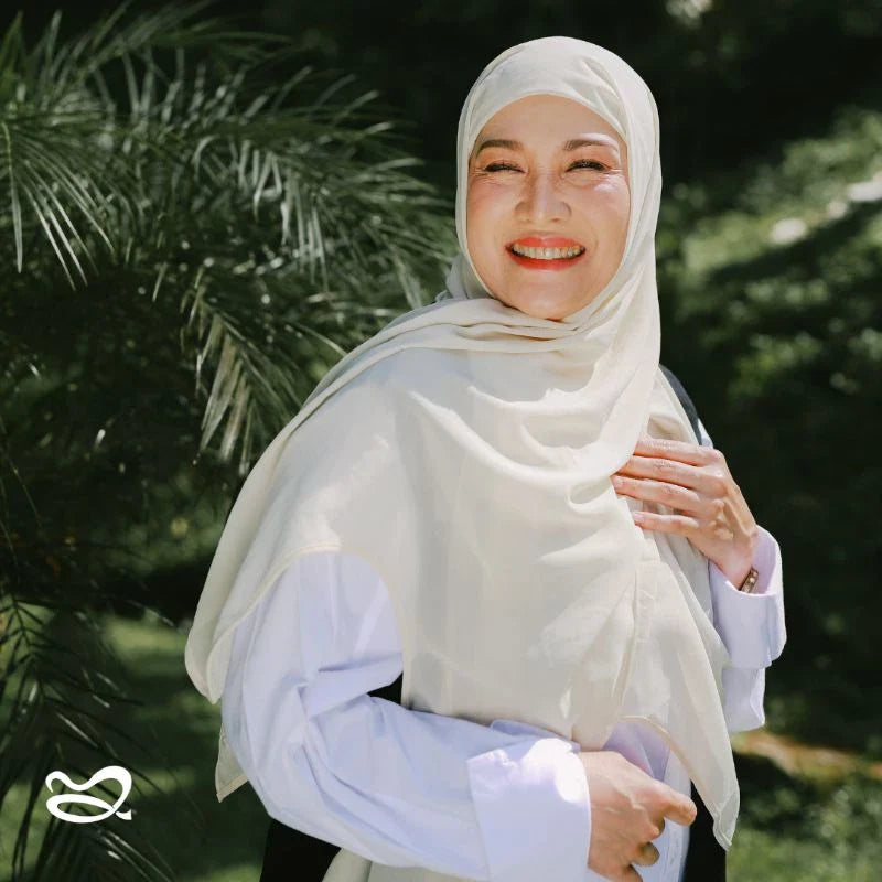 A woman wearing a light yellow pashmina headscarf over a black outfit against a grey background.