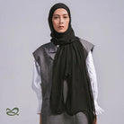 A woman wearing a black pashmina headscarf over a black outfit against a grey background.
