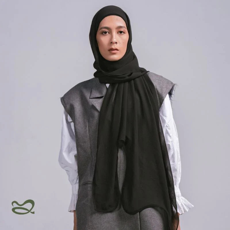 A woman wearing a black pashmina headscarf over a black outfit against a grey background.