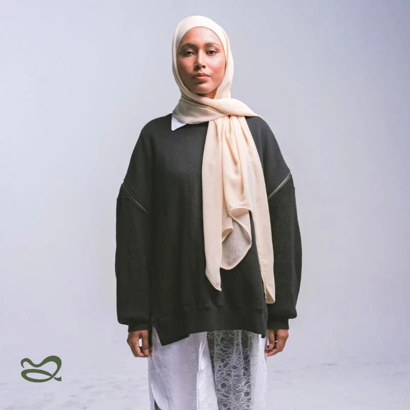 A woman wearing a beige pashmina headscarf over a black outfit against a grey background.