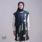 A woman wearing a dark blue pashmina headscarf over a black outfit against a grey background.