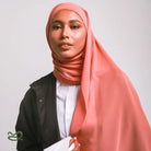 A woman wearing a taupe-colored pashmina headscarf over a black outfit against a grey background.