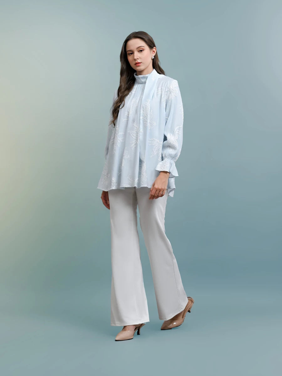 Woman wearing a light blue blouse with floral patterns by Kasa Heritage and white pants against a gray background