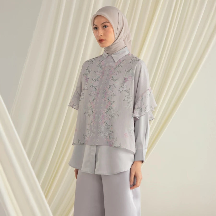 Woman wearing a light gray floral shirt by Hijabchic and pants with a matching hijab against a white curtain background.