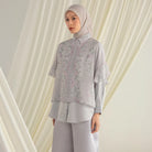 Woman wearing a light gray floral shirt by Hijabchic and pants with a matching hijab against a white curtain background.